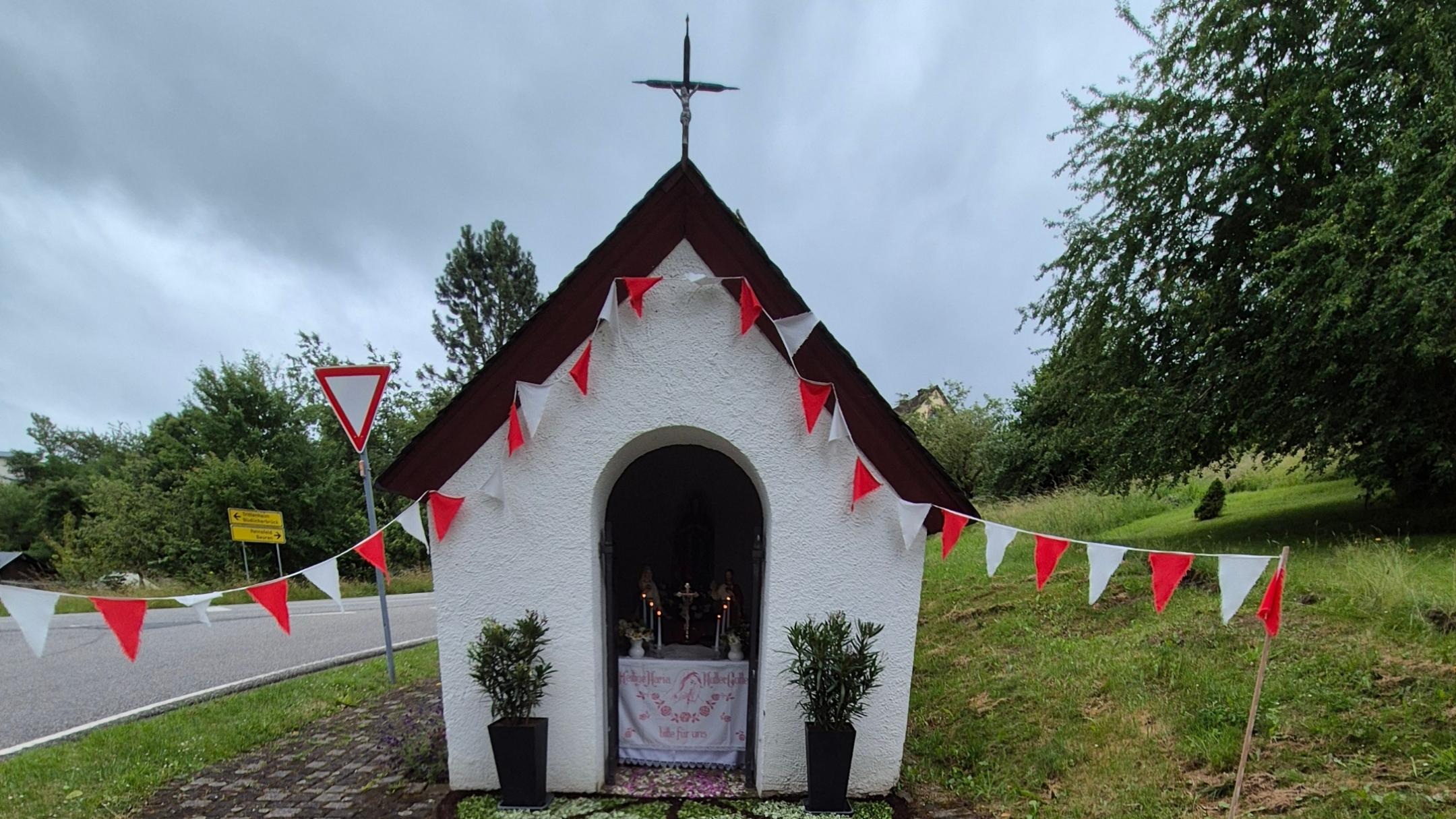 Kleine weiße Kapelle mit Kreuz auf dem Dach, geschmückt mit roten und weißen Wimpeln im Grünen.