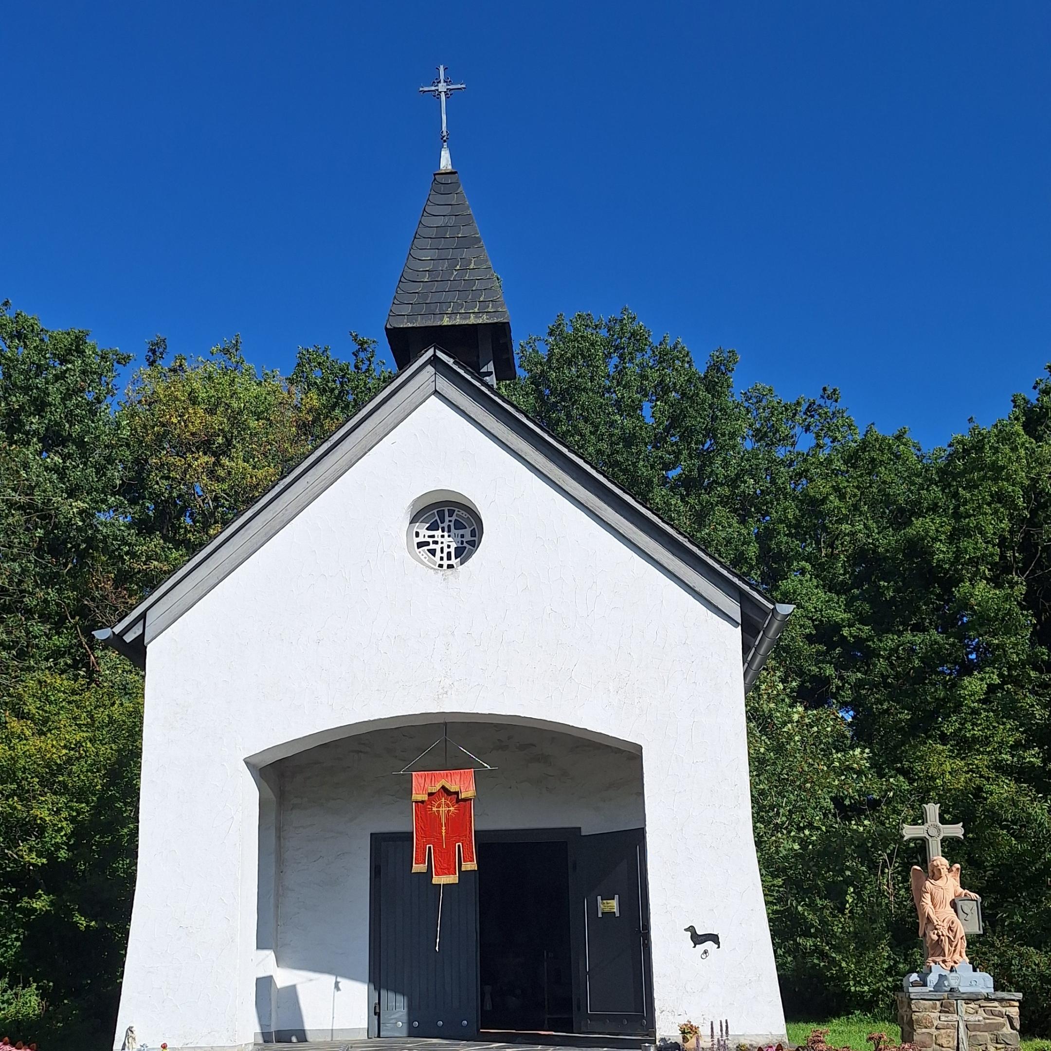 Das Bild zeigt eine kleine weiße Kapelle mit schwarzem Dach und Turmkreuz, einem roten Banner über dem Eingang, einer Rampe an der Seite und einer Jesusstatue am Kreuz im Vordergrund, umgeben von Bäumen und Grün.