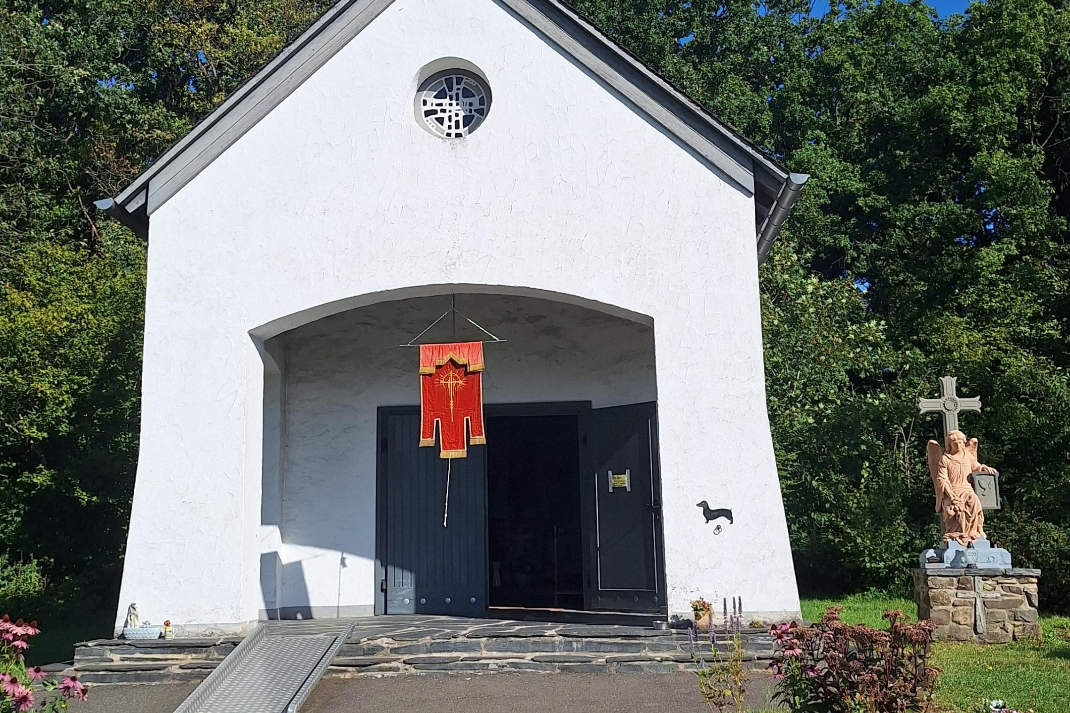 Das Bild zeigt eine kleine weiße Kapelle mit schwarzem Dach und Turmkreuz, einem roten Banner über dem Eingang, einer Rampe an der Seite und einer Jesusstatue am Kreuz im Vordergrund, umgeben von Bäumen und Grün.