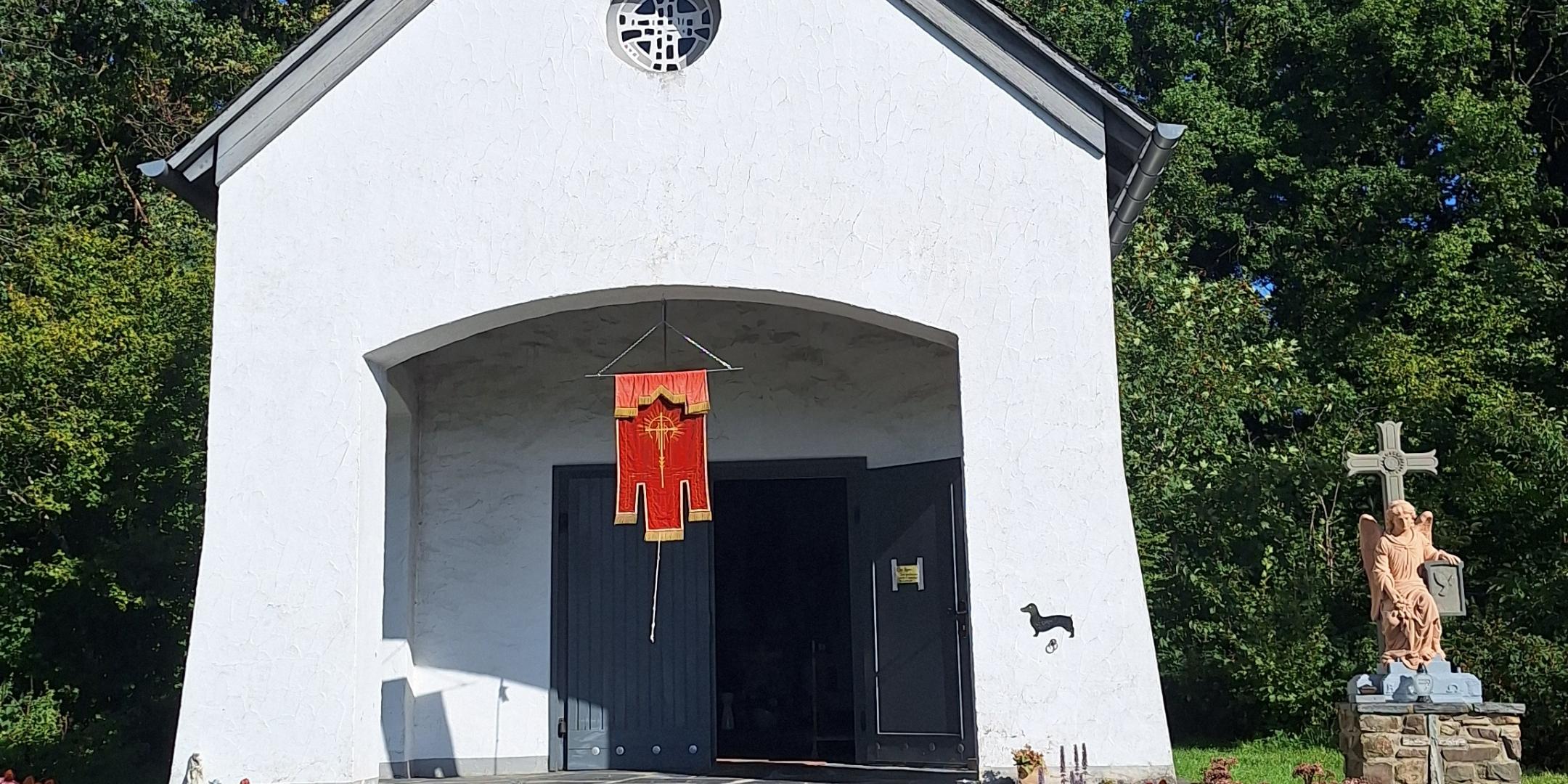 Das Bild zeigt eine kleine weiße Kapelle mit schwarzem Dach und Turmkreuz, einem roten Banner über dem Eingang, einer Rampe an der Seite und einer Jesusstatue am Kreuz im Vordergrund, umgeben von Bäumen und Grün.