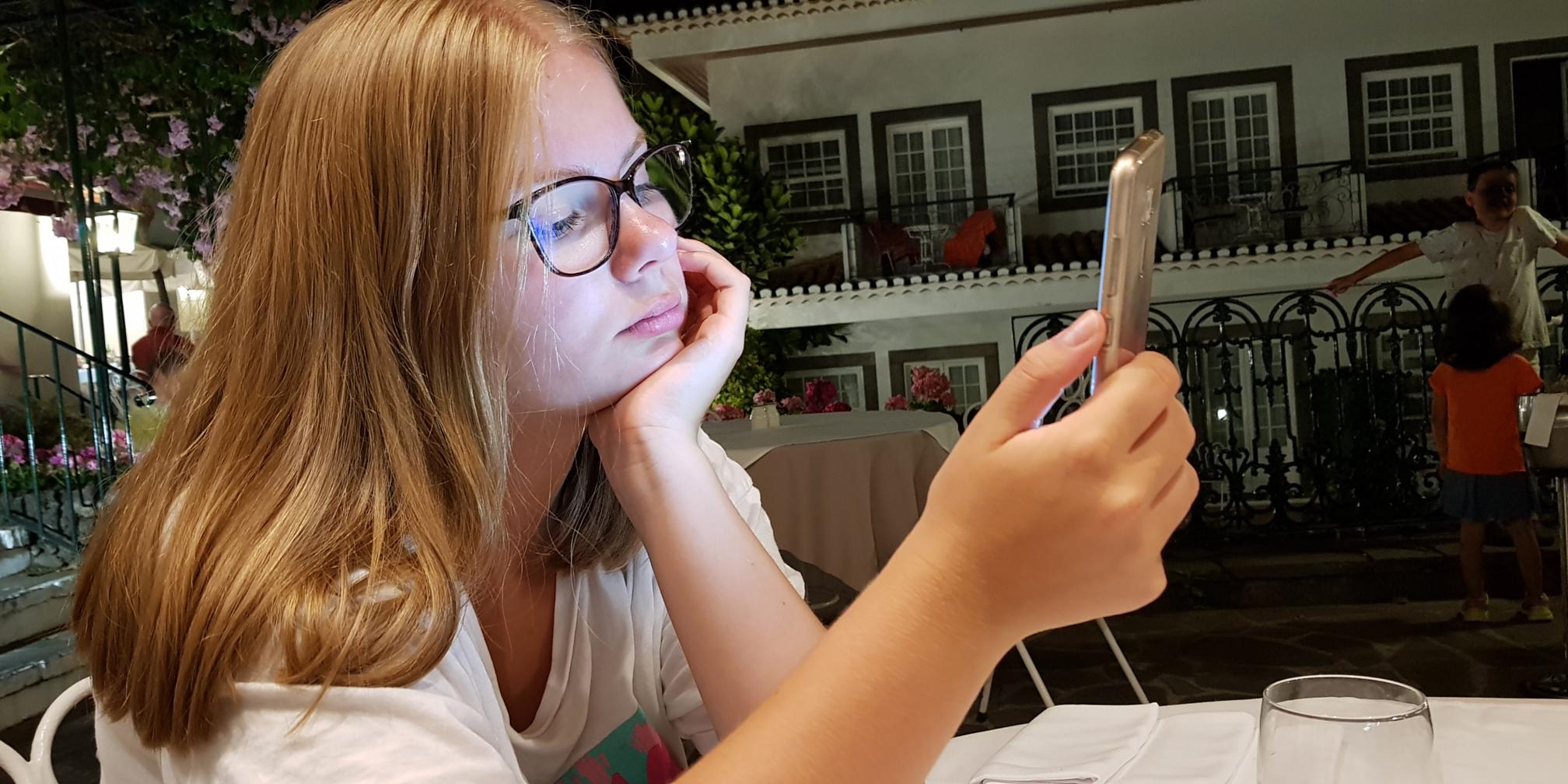 Das Foto zeigt ein Mädchen, das an einem Tisch im Freien sitzt und ein Smartphone hält, während im Hintergrund ein Gebäude mit Balkonen und weitere Menschen zu sehen sind.