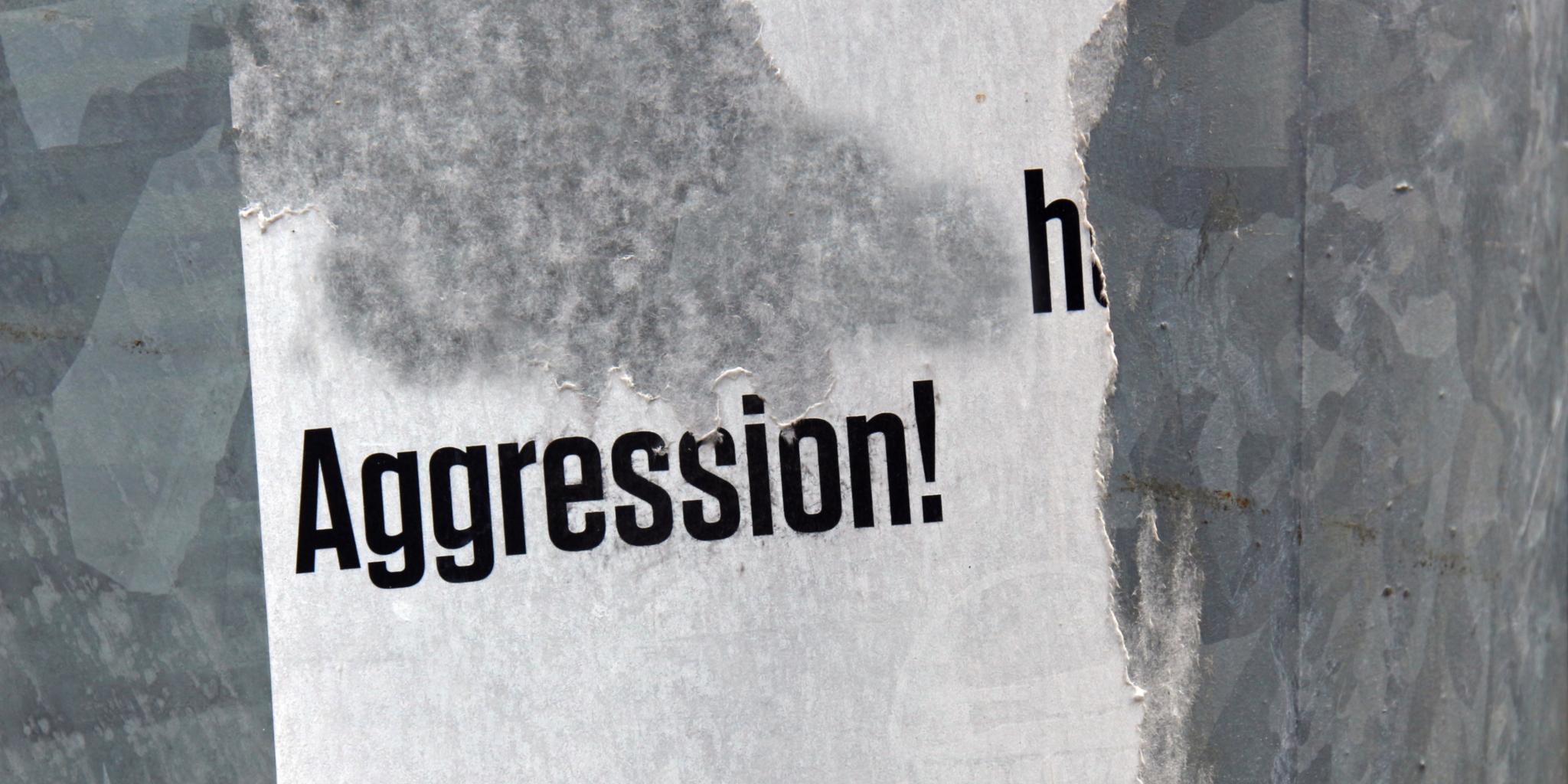 Das Foto zeigt ein teilweise abgerissenes Plakat mit dem Wort „Aggression!“ in schwarzer Schrift auf weißem Hintergrund.