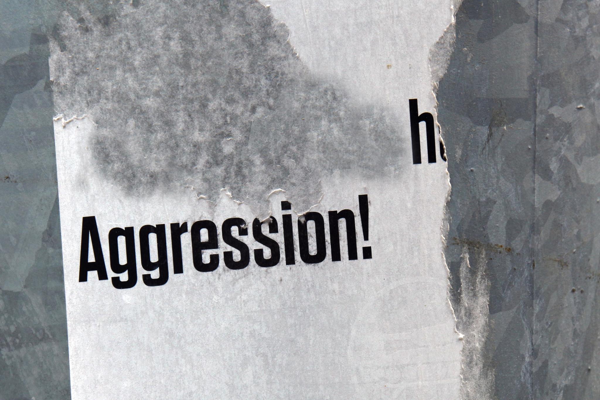 Das Foto zeigt ein teilweise abgerissenes Plakat mit dem Wort „Aggression!“ in schwarzer Schrift auf weißem Hintergrund.