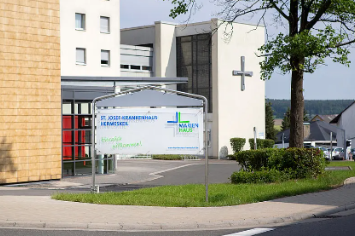 Das Foto zeigt den sonnigen Eingangsbereich eines modernen Krankenhauses mit einem Willkommensschild vor einer Fassade aus gelben Ziegeln und einem weißen Gebäudeteil mit großem Kreuz.