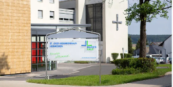 Das Foto zeigt den sonnigen Eingangsbereich eines modernen Krankenhauses mit einem Willkommensschild vor einer Fassade aus gelben Ziegeln und einem weißen Gebäudeteil mit großem Kreuz.