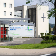 Das Foto zeigt den sonnigen Eingangsbereich eines modernen Krankenhauses mit einem Willkommensschild vor einer Fassade aus gelben Ziegeln und einem weißen Gebäudeteil mit großem Kreuz.