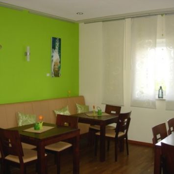 Ein kleiner, gemütlicher Raum mit grüner Akzentwand, schlichten Holztischen und einer Sitzbank, dekoriert mit Pflanzen und hellen Vorhängen. (c) Team Mehrgenerationenhaus Ein kleiner, gemütlicher Raum mit grüner Akzentwand, schlichten Holztischen und einer Sitzbank, dekoriert mit Pflanzen und hellen Vorhängen.