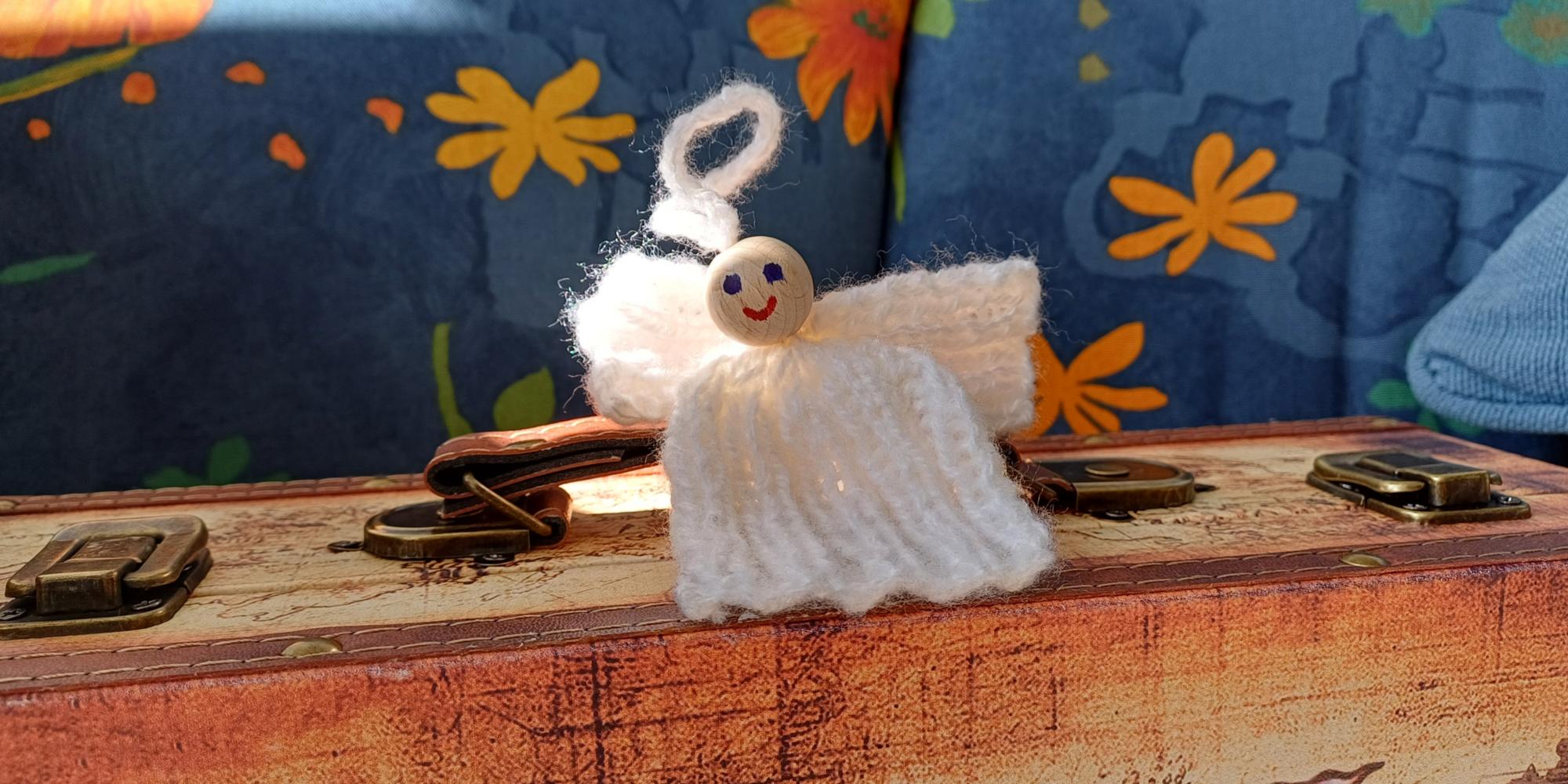 Eine kleine Engelsfigur aus weißem Garn mit einem lächelnden Holzperlenkopf sitzt auf einem alten Koffer, vor einem Hintergrund aus blauem Stoff mit orange-gelben Blumenmustern.
