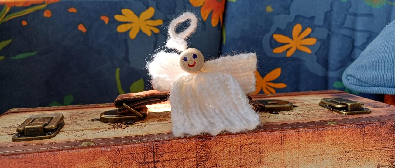 Eine kleine Engelsfigur aus weißem Garn mit einem lächelnden Holzperlenkopf sitzt auf einem alten Koffer, vor einem Hintergrund aus blauem Stoff mit orange-gelben Blumenmustern.