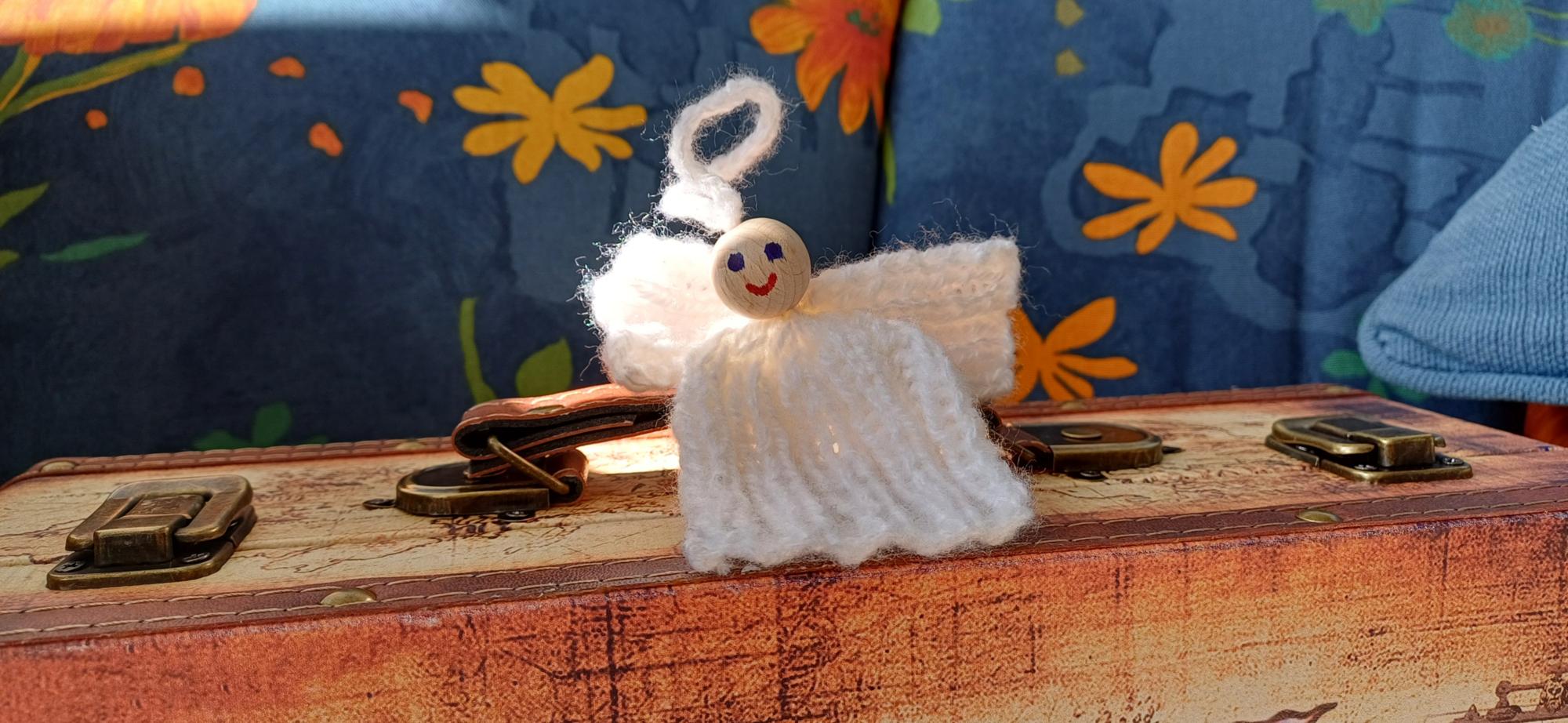 Eine kleine Engelsfigur aus weißem Garn mit einem lächelnden Holzperlenkopf sitzt auf einem alten Koffer, vor einem Hintergrund aus blauem Stoff mit orange-gelben Blumenmustern.