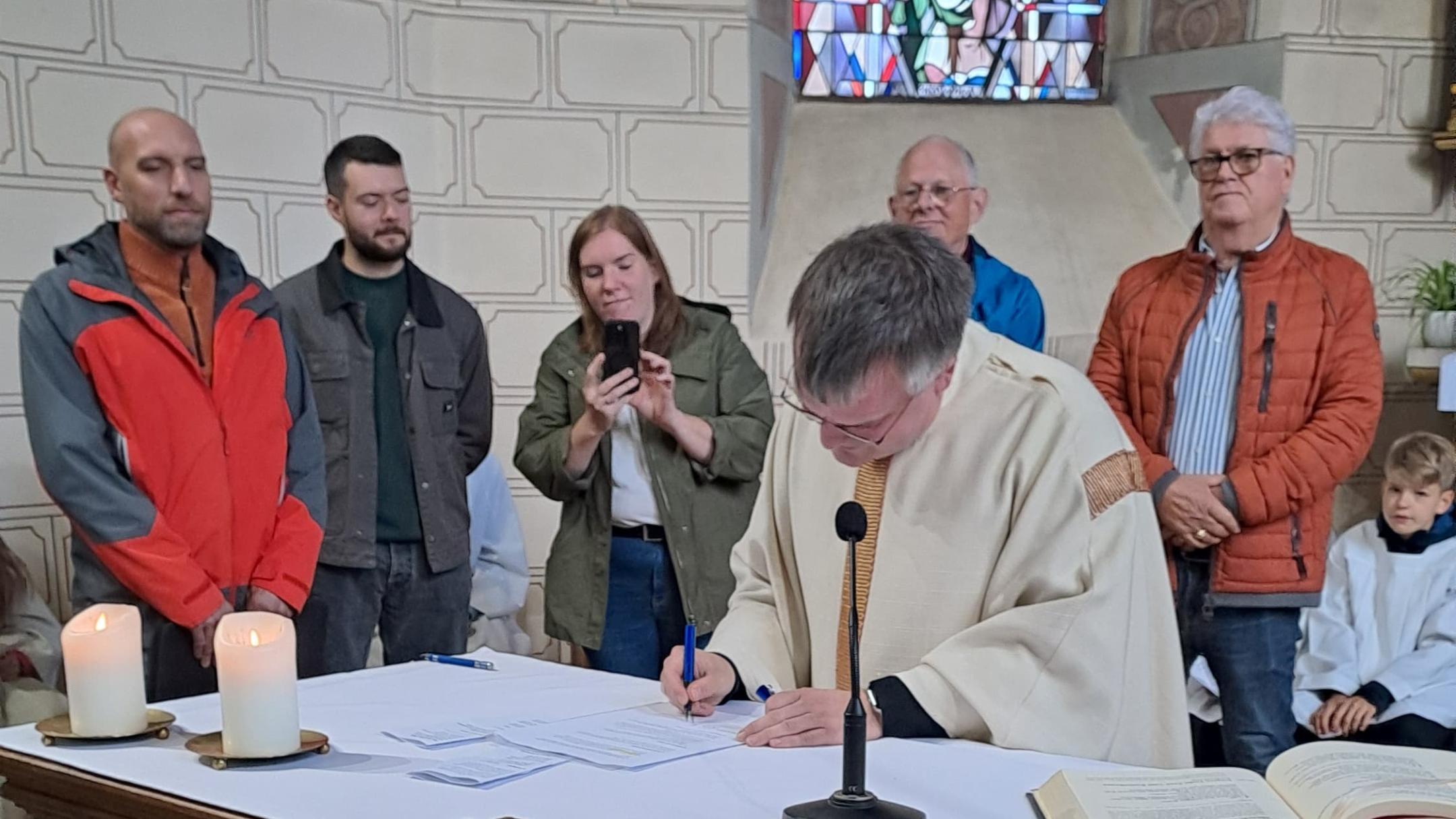 Pfarrer Heinz unterschreibt ein Dokument auf dem Altar einer Kirche, umgeben von mehreren Personen, während im Vordergrund Körbe mit Obst und Gemüse und im Hintergrund ein buntes Kirchenfenster zu sehen sind.