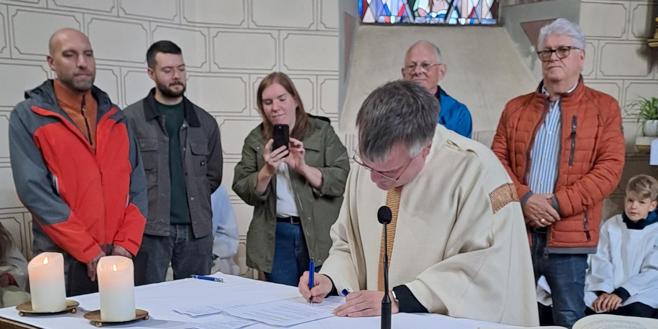 Pfarrer Heinz unterschreibt ein Dokument auf dem Altar einer Kirche, umgeben von mehreren Personen, während im Vordergrund Körbe mit Obst und Gemüse und im Hintergrund ein buntes Kirchenfenster zu sehen sind.