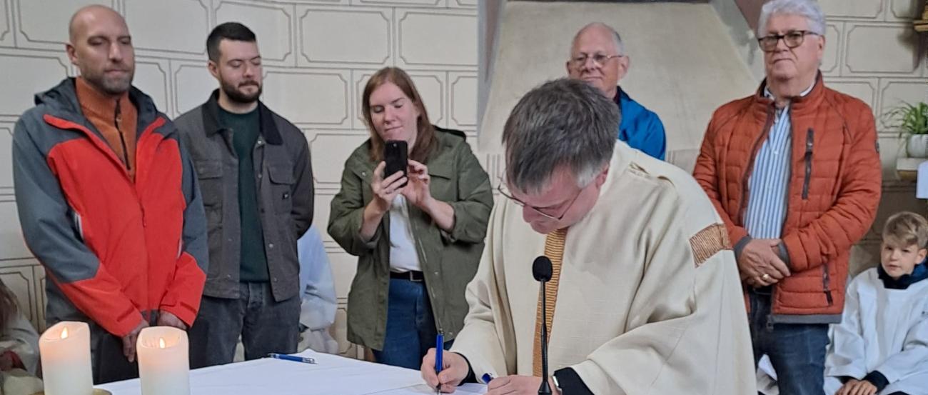 Pfarrer Heinz unterschreibt ein Dokument auf dem Altar einer Kirche, umgeben von mehreren Personen, während im Vordergrund Körbe mit Obst und Gemüse und im Hintergrund ein buntes Kirchenfenster zu sehen sind.
