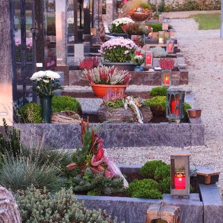 Das Bild zeigt einen Friedhof mit mehreren Gräbern, die liebevoll mit Blumen, Pflanzen und Laternen geschmückt sind.
