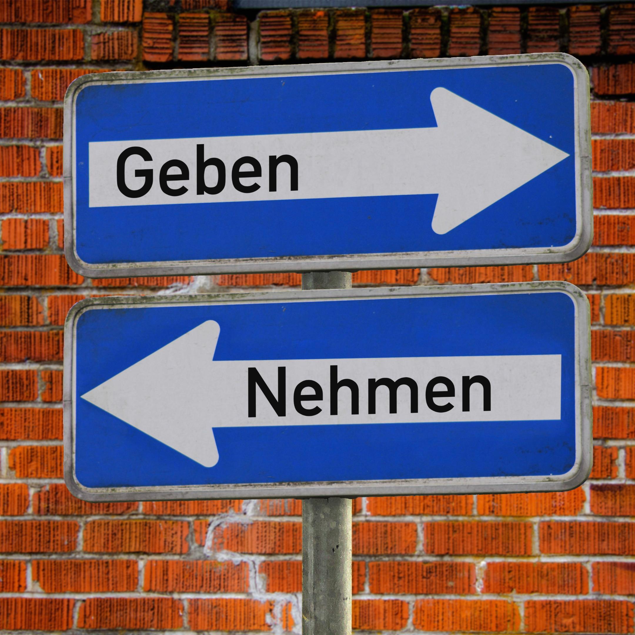 Das Foto zeigt zwei blaue Verkehrsschilder vor einer Backsteinmauer – eines mit einem weißen Pfeil nach rechts und dem Wort „Geben“, das andere mit einem weißen Pfeil nach links und dem Wort „Nehmen“.