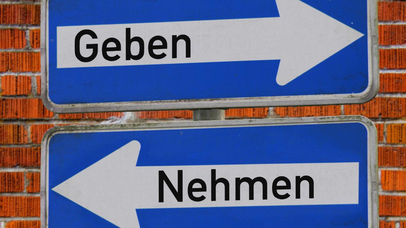 Das Foto zeigt zwei blaue Verkehrsschilder vor einer Backsteinmauer – eines mit einem weißen Pfeil nach rechts und dem Wort „Geben“, das andere mit einem weißen Pfeil nach links und dem Wort „Nehmen“.