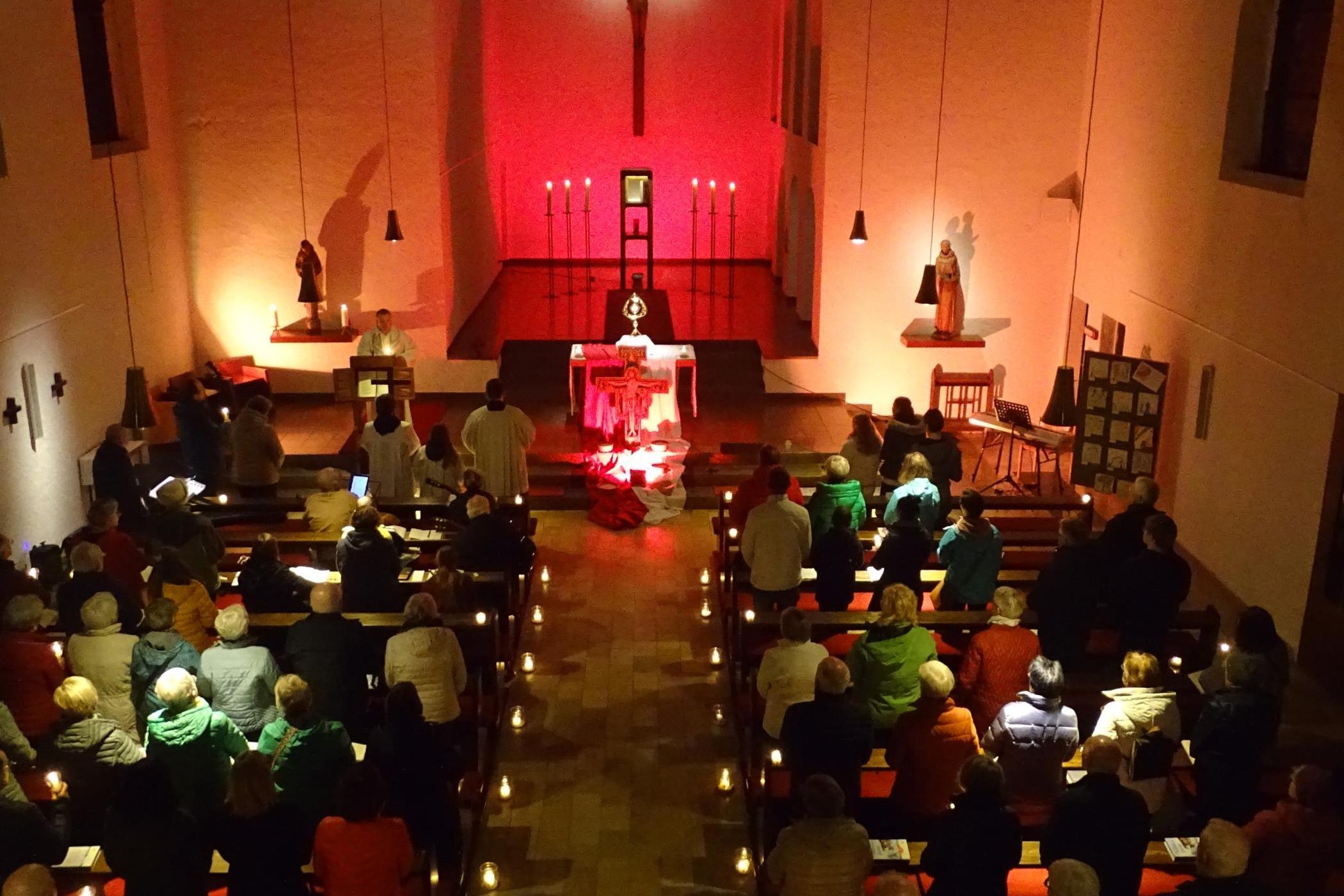 Eine Kerzenandacht in einer Kirche mit rot beleuchtetem Altar und vielen Menschen in den Bänken.