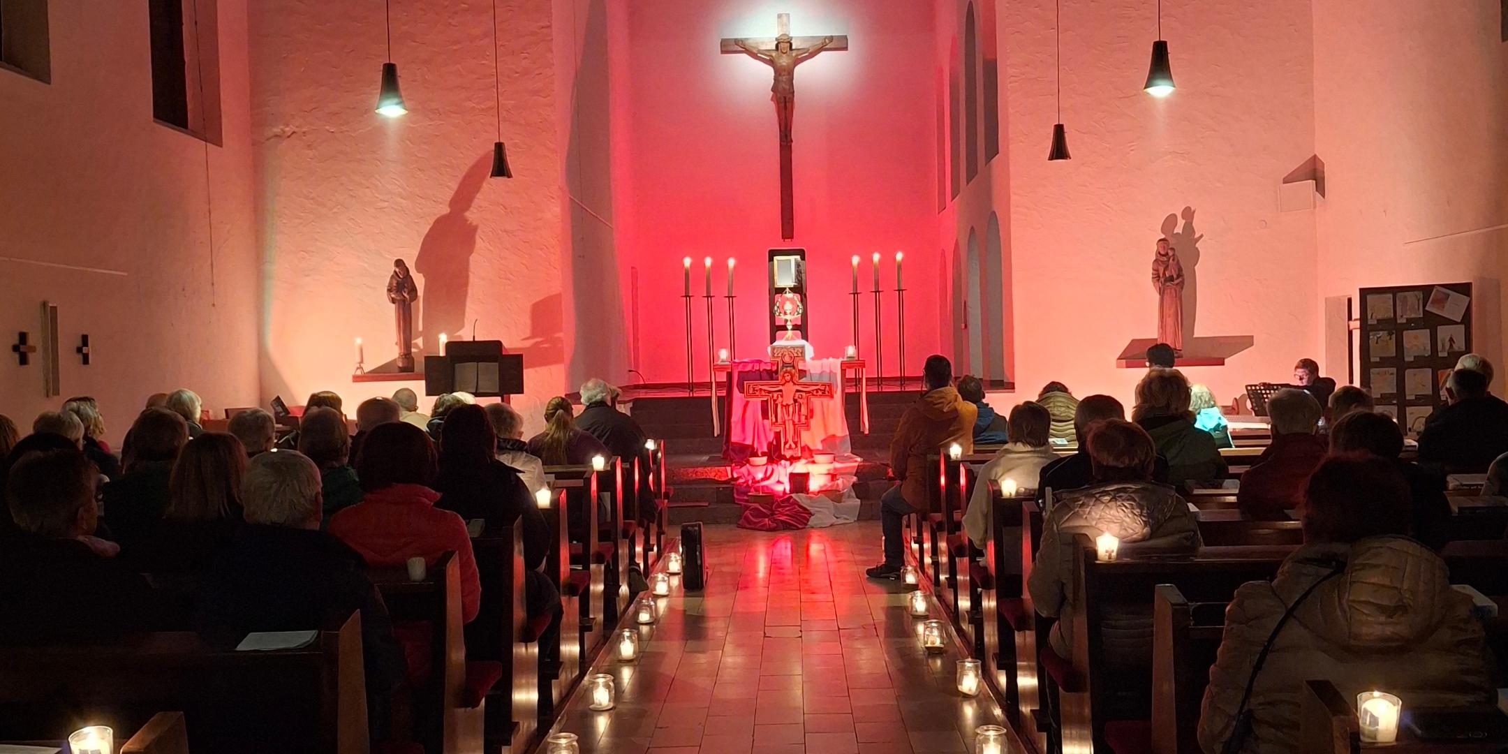 Kirchenraum mit Kerzenlicht und Gottesdienstbesuchern vor einem rot beleuchteten Altar.