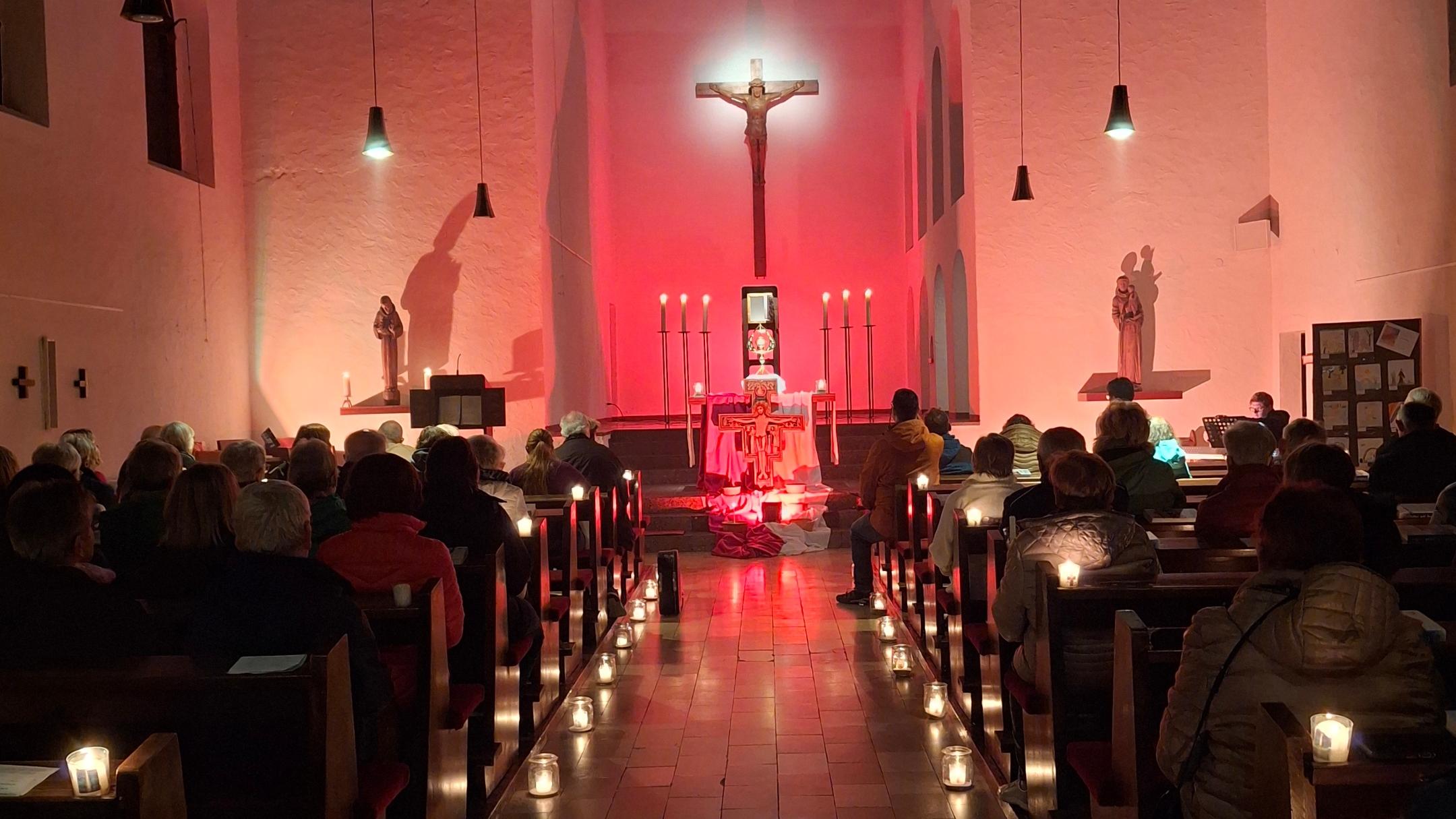 Kirchenraum mit Kerzenlicht und Gottesdienstbesuchern vor einem rot beleuchteten Altar.