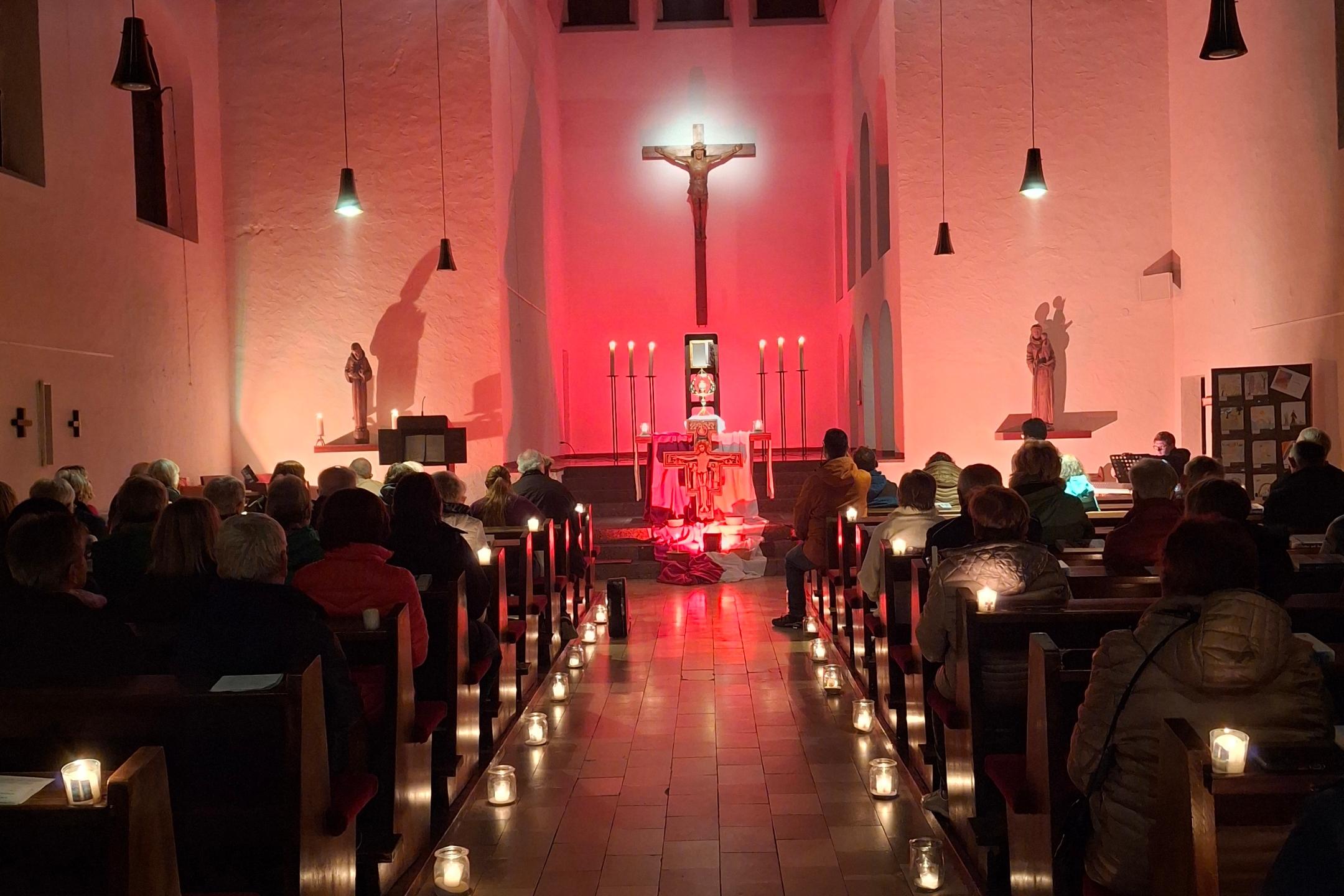 Kirchenraum mit Kerzenlicht und Gottesdienstbesuchern vor einem rot beleuchteten Altar.