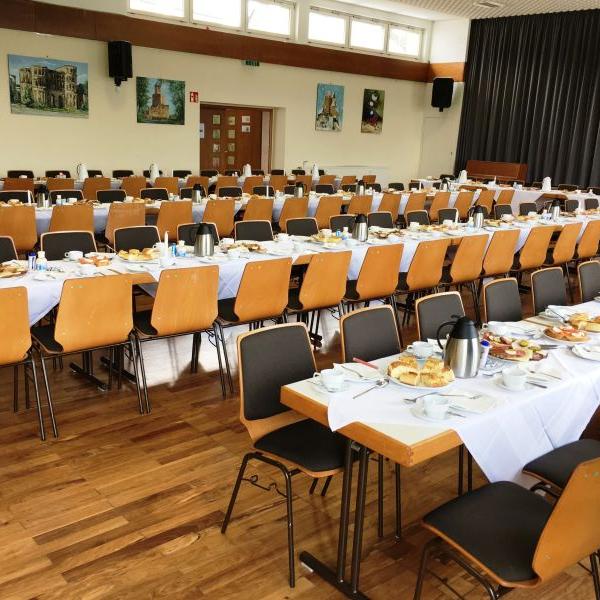 Ein großer Saal ist mit langen, festlich gedeckten Tischen voller Geschirr, Thermoskannen und Speisen vorbereitet. (c) Team Mehrgenerationenhaus Ein großer Saal ist mit langen, festlich gedeckten Tischen voller Geschirr, Thermoskannen und Speisen vorbereitet.