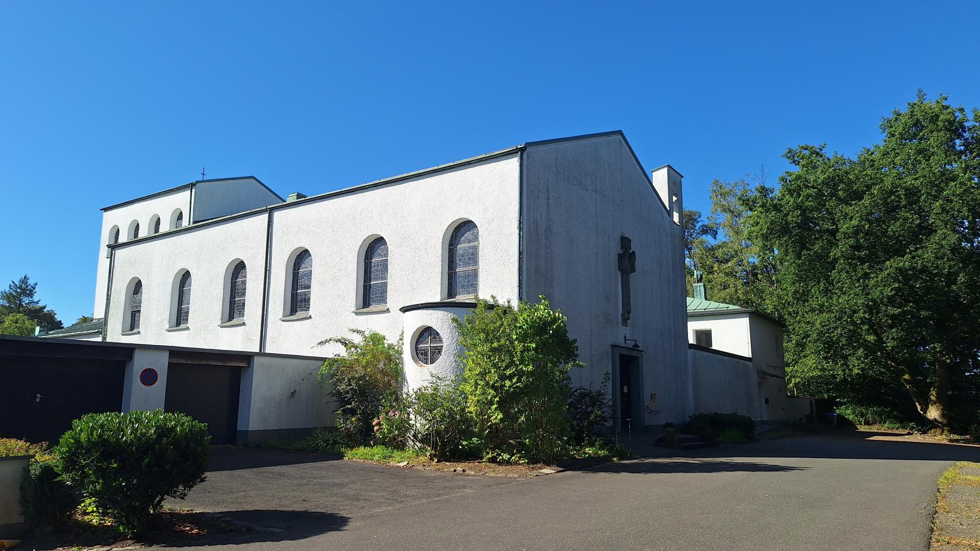 Das Bild zeigt eine weiße Kirche mit Rundbogenfenstern und einem Kreuz an der Fassade, sie steht umgeben von Bäumen und Grünflächen. Der Himmel ist klar und blau, was der Szene eine ruhige, friedliche Stimmung verleiht.