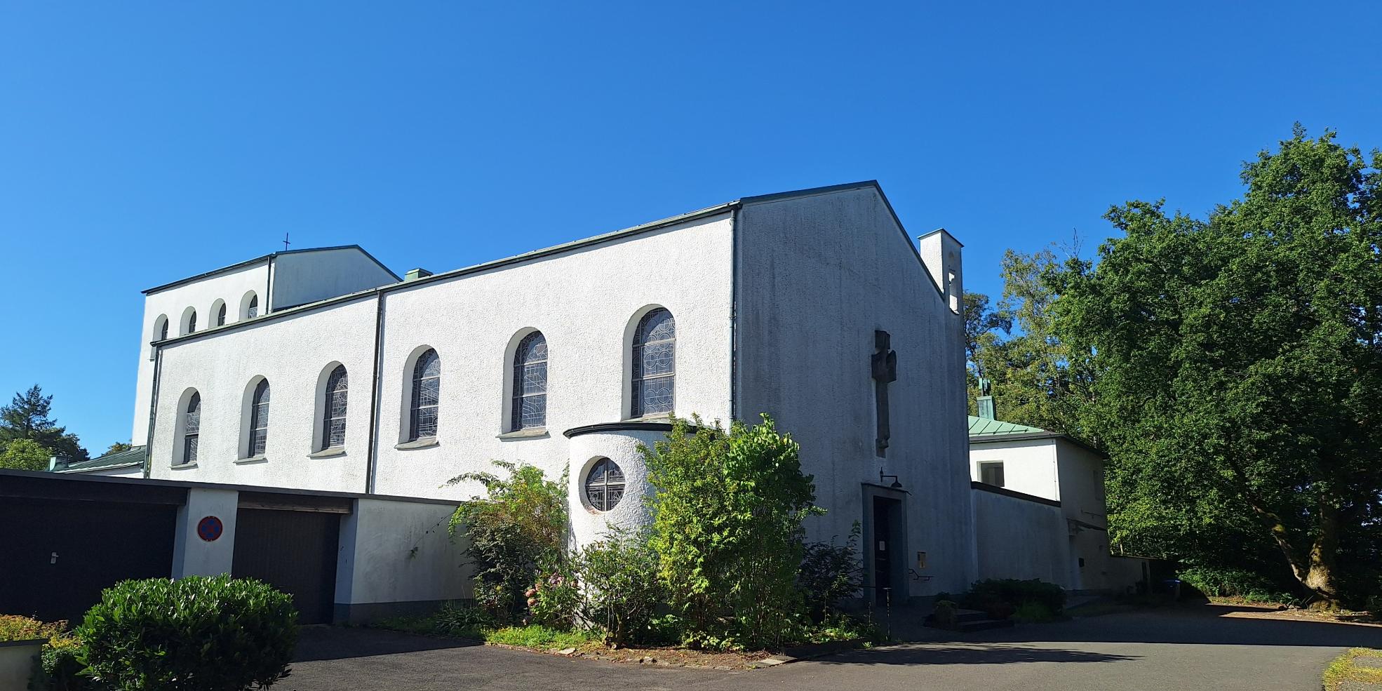 Das Bild zeigt eine weiße Kirche mit Rundbogenfenstern und einem Kreuz an der Fassade, sie steht umgeben von Bäumen und Grünflächen. Der Himmel ist klar und blau, was der Szene eine ruhige, friedliche Stimmung verleiht.