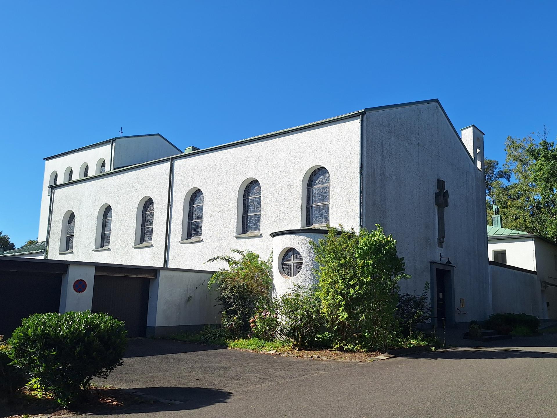 Das Bild zeigt eine weiße Kirche mit Rundbogenfenstern und einem Kreuz an der Fassade, sie steht umgeben von Bäumen und Grünflächen. Der Himmel ist klar und blau, was der Szene eine ruhige, friedliche Stimmung verleiht.