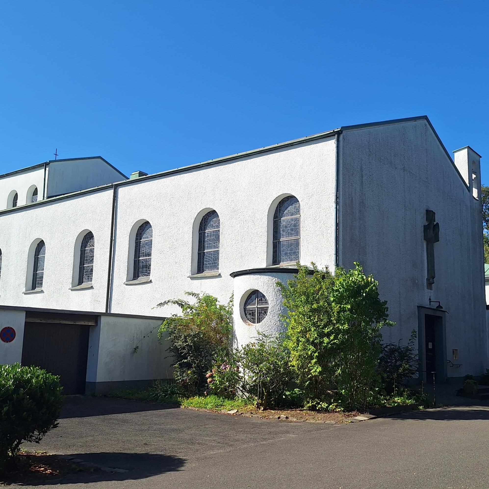 Das Bild zeigt eine weiße Kirche mit Rundbogenfenstern und einem Kreuz an der Fassade, sie steht umgeben von Bäumen und Grünflächen. Der Himmel ist klar und blau, was der Szene eine ruhige, friedliche Stimmung verleiht.