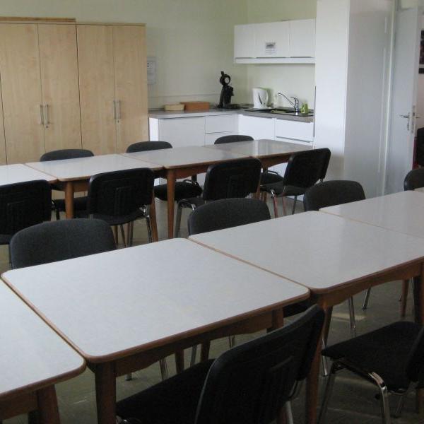 Ein schlichter Mehrzweckraum mit hellen Tischen, schwarzen Stühlen und einer kleinen Küchenzeile. (c) Team Mehrgenerationenhaus Ein schlichter Mehrzweckraum mit hellen Tischen, schwarzen Stühlen und einer kleinen Küchenzeile.