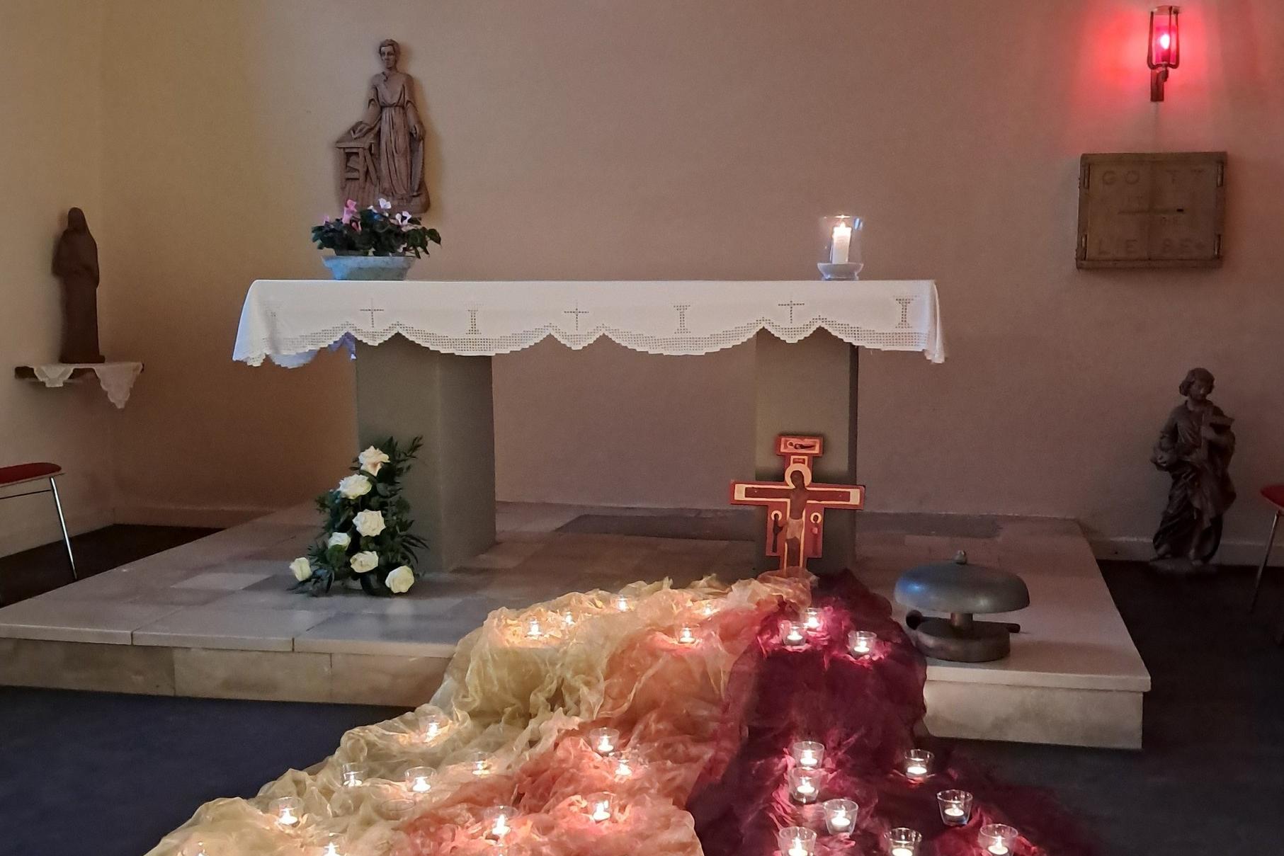 Auf dem Bild ist ein schlichter Kirchenaltar zu sehen, vor dem farbige Tücher mit vielen kleinen brennenden Kerzen liegen, darüber ein Kreuz