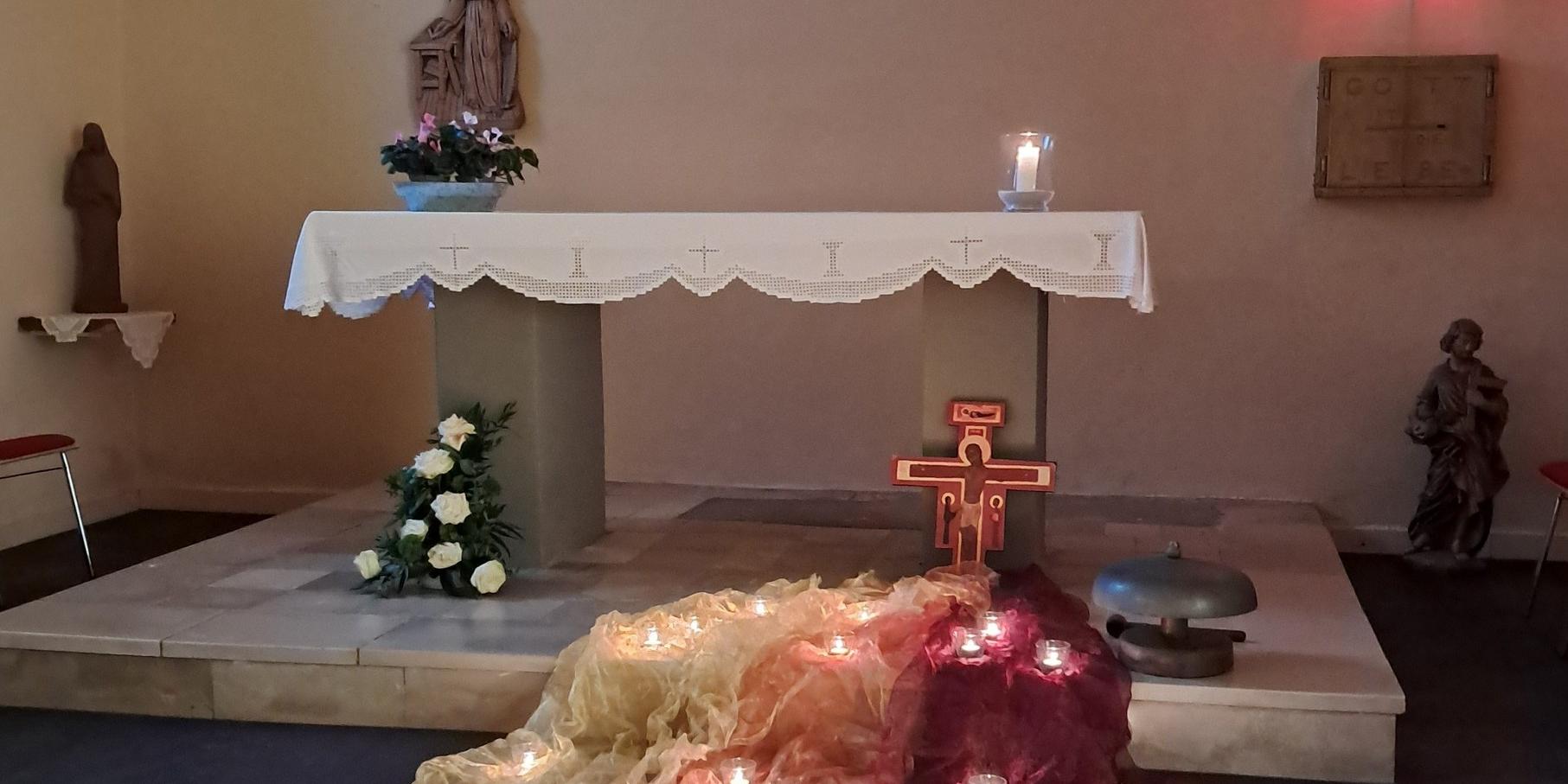 Auf dem Bild ist ein schlichter Kirchenaltar zu sehen, vor dem farbige Tücher mit vielen kleinen brennenden Kerzen liegen, darüber ein Kreuz