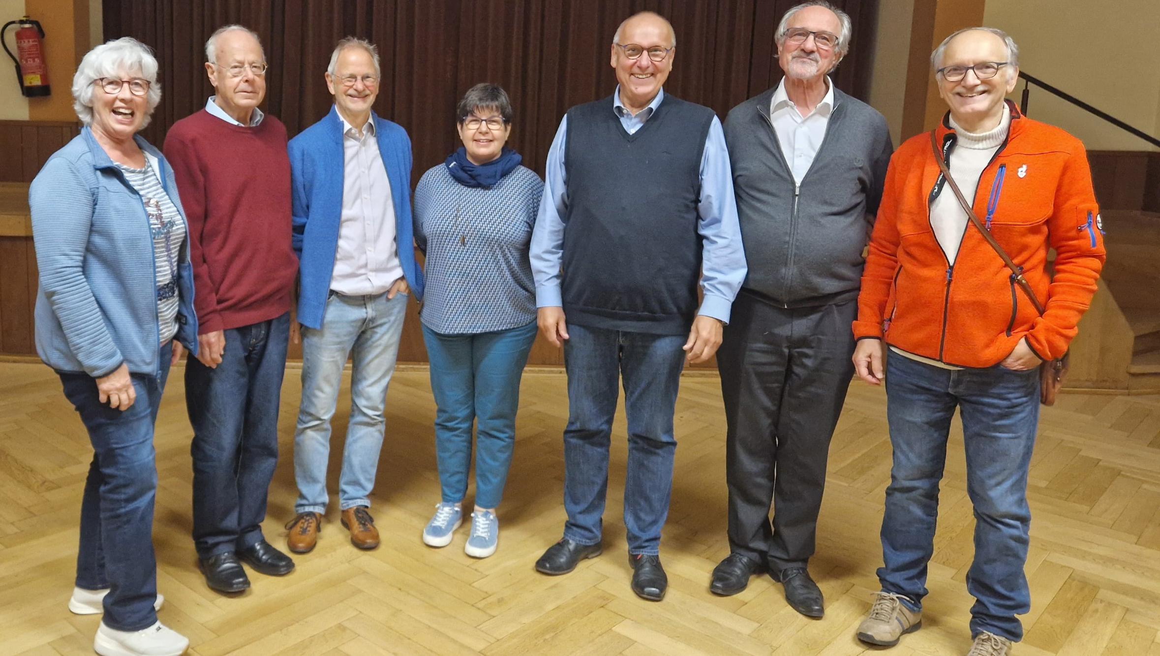 Sieben erwachsene Personen stehen nebeneinander in einem Raum mit Holzfußboden und Vorhang im Hintergrund.