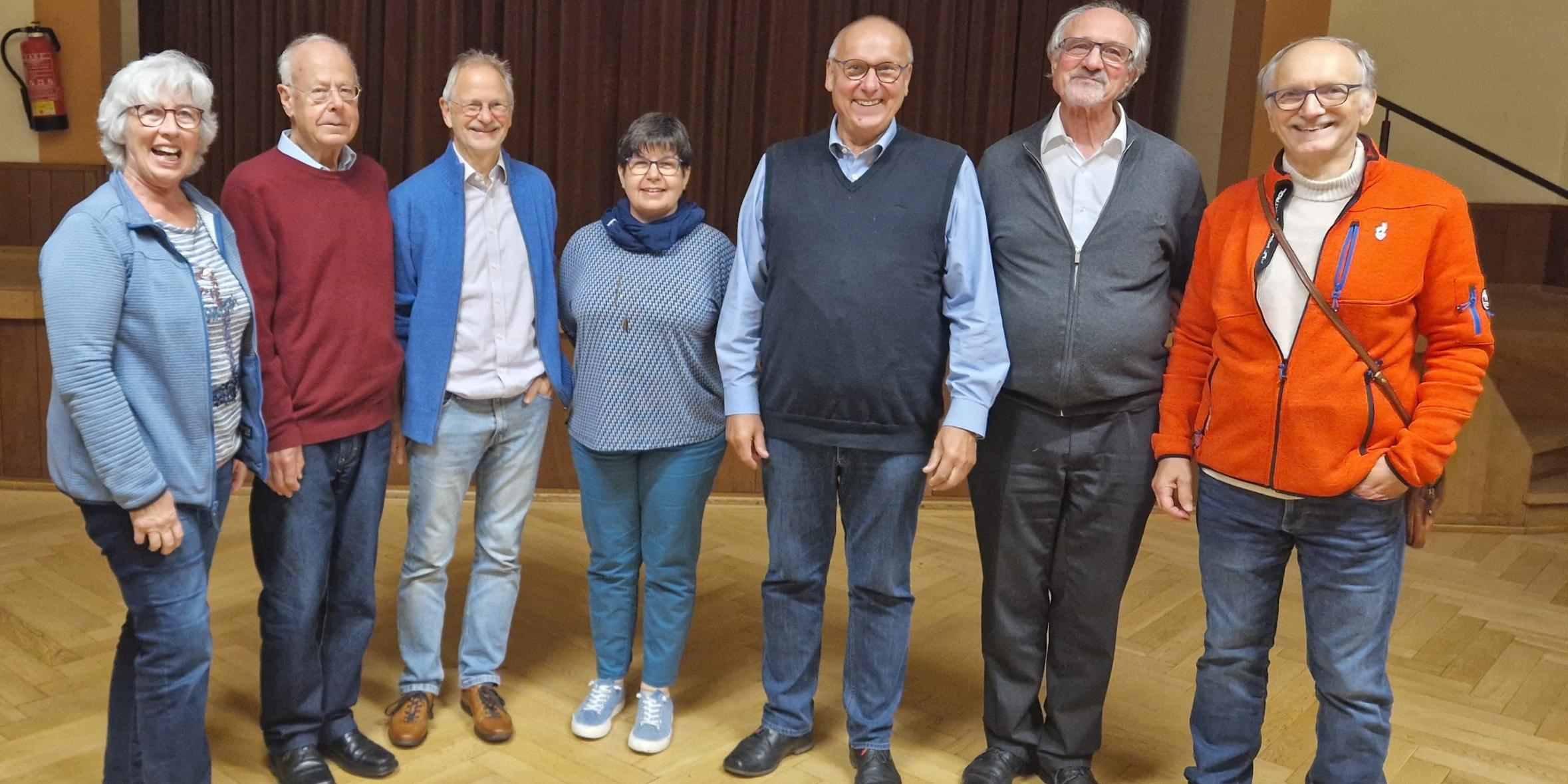 Sieben erwachsene Personen stehen nebeneinander in einem Raum mit Holzfußboden und Vorhang im Hintergrund.