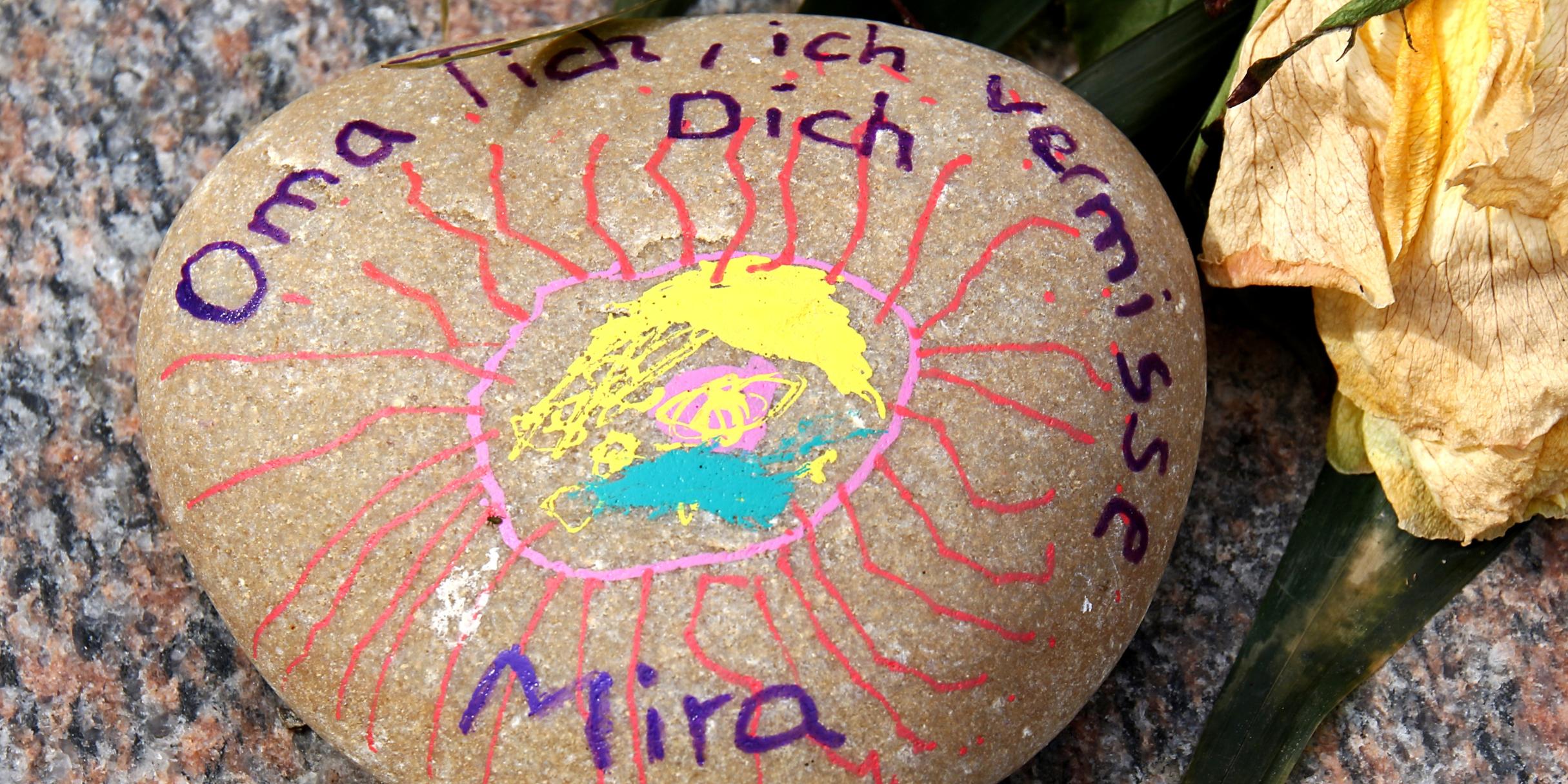 Ein bemalter Stein mit einer gezeichneten Sonne und der Aufschrift „Oma ich vermisse Dich – Mira“ liegt neben einer verwelkten gelben Blume auf einer Oberfläche.