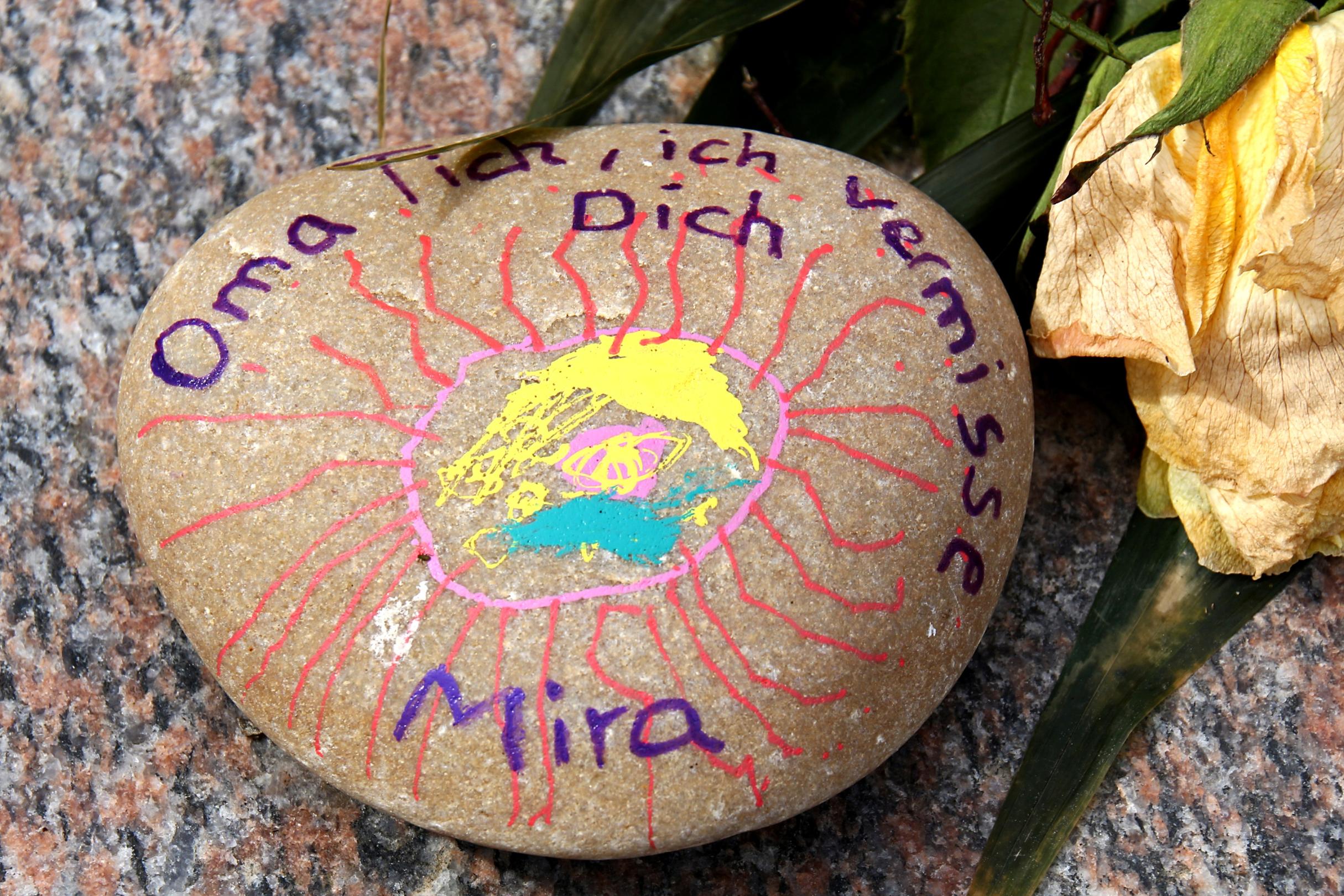 Ein bemalter Stein mit einer gezeichneten Sonne und der Aufschrift „Oma ich vermisse Dich – Mira“ liegt neben einer verwelkten gelben Blume auf einer Oberfläche.