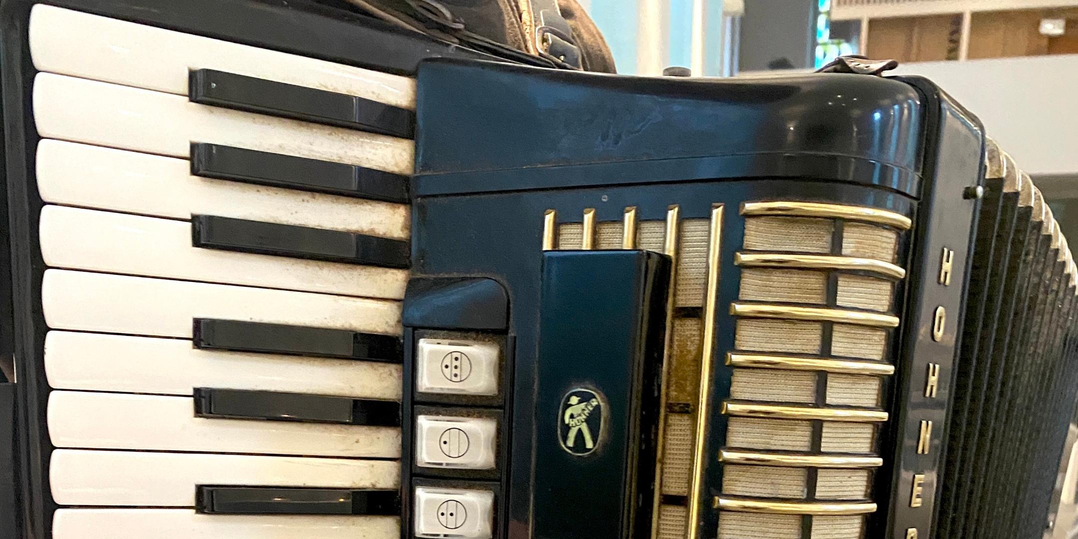 Das Bild zeigt eine Person, die ein schwarzes Akkordeon der Marke „Hohner“ spielt – mit weißen Tasten auf der linken und goldenen Knöpfen auf der rechten Seite