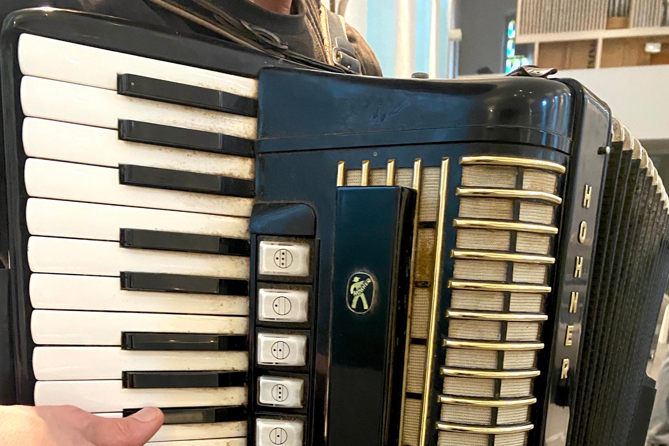 Das Bild zeigt eine Person, die ein schwarzes Akkordeon der Marke „Hohner“ spielt – mit weißen Tasten auf der linken und goldenen Knöpfen auf der rechten Seite