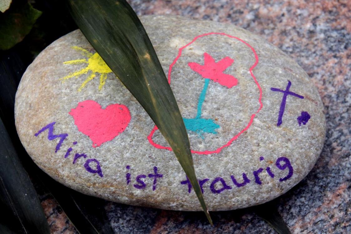 Ein bemalter Stein liegt auf einem größeren Stein, darauf sind eine gelbe Sonne, ein rotes Herz, eine rote Blume mit grünem Stiel, ein lilafarbenes Kreuz und der handgeschriebene Satz „Mira ist traurig“.